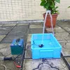 A small water pump is set up in a blue plastic tub, creating a fountain that sprays water upwards. A green battery pack is connected to the pump, and the setup is placed on a tiled outdoor surface next to a potted plant. Podgląd zdjęcia produktu