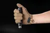 A hand wearing a tan tactical glove holds a black flashlight against a black background. Produktbild-Vorschau