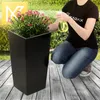 A person is watering a plant in a black planter on a wooden deck. Anteprima dell’immagine prodotto