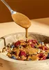A close-up shot of a wooden bowl filled with granola, berries, and nuts, with almond butter being drizzled from a spoon. Produktbild-Vorschau