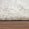 A close-up view of a fluffy white rug on a wooden floor. Produktbild-Vorschau