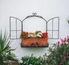 A decorative metal window frame with a planter box filled with artificial flowers mounted on a white wall. Produktbild-Vorschau