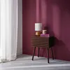 A bedside table with a lamp and books next to a window with sheer curtains. Produktbild-Vorschau