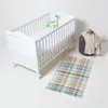 A white baby crib with a green dinosaur toy on the mattress, a grey ottoman with blankets, and a plaid rug on a white floor. Produktbild-Vorschau