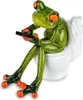 A decorative figurine of a frog sitting on a toilet and holding a remote control. Produktbild-Vorschau