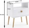 A white bedside table with wooden legs and a drawer with a wooden knob, shown with dimensions indicated by text and lines. Podgląd zdjęcia produktu