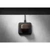 A brown leather case for AirPods is placed on a black wireless charging pad, which is connected to a cable and mounted on a textured gray wall. Podgląd zdjęcia produktu
