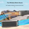 Two black Bluetooth speakers are shown on a sandy surface with rocks and a blurred backpack in the background. Blue and orange light effects emanate from the speakers, suggesting sound waves. Podgląd zdjęcia produktu