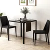 A black glass-top dining table with two black chairs in a room with light wood flooring and framed pictures on the wall. Produktbild-Vorschau