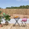 An outdoor patio scene with a bamboo fence, plants, a table, and a chair. Anteprima dell’immagine prodotto