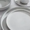 A close-up view of speckled white ceramic plates and bowls stacked on a light gray textured surface. Produktbild-Vorschau