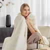 A woman is sitting on a chair, wrapped in a soft, off-white blanket. She is looking away from the camera with a relaxed expression. The setting appears to be a living room with a vase of dried flowers and a cabinet in the background. Produktbild-Vorschau