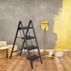 A black metal step ladder is set up in a room with a gray wall being painted yellow. There are cardboard boxes and paint cans nearby. Produktbild-Vorschau