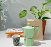 A green Bialetti Moka pot sits on a table next to a small patterned cup, a stack of books, and a potted plant with variegated leaves. Podgląd zdjęcia produktu
