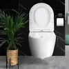 A modern white wall-mounted toilet with the lid up, set against a black marble tiled wall and gray tiled floor. A potted plant with green leaves is to the left, and a toilet paper holder with a roll is to the right. Produktbild-Vorschau