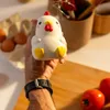A hand holding a chicken-shaped stress toy in a kitchen setting with eggs and tomatoes visible. Voir les images du produit