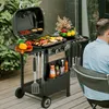 A person is sitting at a table next to a gas grill and griddle combo, which is loaded with food. Produktbild-Vorschau