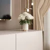 A close-up view of a white vase with white roses on top of a white cabinet, with curtains and decorative lighting in the background. Náhled obrázku produktu