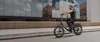 A man rides an electric bicycle on a city street with modern buildings in the background. Produktbild-Vorschau