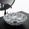 A decorative ceramic washbasin with a faucet and mirror in the background, placed on a black countertop. Náhled obrázku produktu