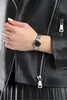 A close-up shot of a woman's wrist wearing a silver watch with a black face, layered over a black leather jacket. Produktbild-Vorschau