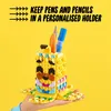 A hand holding a yellow LEGO DOTS pencil holder shaped like a banana, with pens and pencils inside. The background is bright yellow. Podgląd zdjęcia produktu