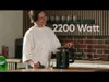 A woman is in a kitchen, standing next to a black electric kettle. There are glasses on the counter, and the text '2200 Watt' is displayed prominently. Produktbild-Vorschau
