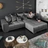 A woman is sitting on a large gray sectional sofa in a modern living room. There are two coffee tables in front of the sofa, and a large abstract chandelier hangs from the ceiling. Produktbild-Vorschau