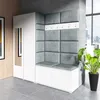 A modern hallway or entrance area featuring a white wardrobe with a mirror, a corner bench with upholstered gray cushions, and a coat rack above the bench. A large potted plant is visible to the right, and a window with a city view is in the background. Produktbild-Vorschau