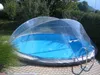 A round swimming pool covered by a clear dome structure, set in a garden with paved ground and trees in the background. Náhľad obrázku produktu