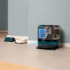 A robot vacuum cleaner with its docking station, shown with internal water tanks and a cutaway view, is positioned on a tiled floor next to two pet bowls. Produktbild-Vorschau
