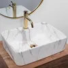 A close-up shot of a rectangular marble-patterned sink with a gold faucet, placed on a wooden surface. A circular mirror is visible in the background. Náhľad obrázku produktu