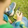 A child's hand holding a yellow and black walkie-talkie, with a blue and green sneaker in the background, on grass. Produktbild-Vorschau
