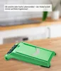 A green vegetable slicer is placed on a wooden table in a kitchen setting. The background is blurred. Voir les images du produit