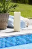A large cream-colored LED candle stands on a wooden deck next to a swimming pool. A potted plant is visible to the left, and a blue blanket and a woven pouf are in the background. Podgląd zdjęcia produktu