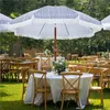 An outdoor wedding or event setup with tables and chairs covered in white tablecloths, set up on a green lawn under striped umbrellas. Produktbild-Vorschau