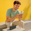 A man is sitting outdoors, smiling while looking at his phone, which is connected to a power bank. Produktbild-Vorschau