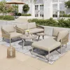 An outdoor patio scene featuring a five-piece garden lounge set, including a sofa, armchair, chaise lounge, and two coffee tables, set on a rug with a backdrop of a building and landscaping. Podgląd zdjęcia produktu