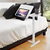 A white adjustable laptop stand is placed next to a bed with white sheets and black pillows. A tablet is on the stand, displaying a landscape image. A gray mug and a stylus are also on the stand. Voir les images du produit
