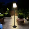 A modern floor lamp illuminates a patio at night, surrounded by lush greenery and outdoor furniture. Voir les images du produit