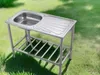 An outdoor stainless steel sink and drainboard on a metal stand, placed on a grassy lawn. Produktbild-Vorschau