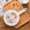 A white handheld coffee or tea strainer with a mesh filter sits on a wooden tray with coffee beans scattered around. Produktbild-Vorschau