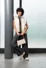A woman leans against a pillar, holding a small handbag. Produktbild-Vorschau