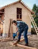 A man is drilling a piece of wood in front of a house under construction. Produktbild-Vorschau