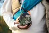 A person is holding a JBL Clip 5 portable speaker with a camouflage pattern. The person is wearing a puffer vest and a fanny pack across their chest. The background is blurred and appears to be outdoors. Produktbild-Vorschau