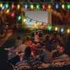 A family is having an outdoor movie night, sitting on blankets and watching a movie projected onto a screen. String lights are hung above them. Produktbild-Vorschau
