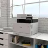 A Canon printer is placed on a white shelf unit next to a window with a brick wall. Produktbild-Vorschau