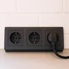 A black triple electrical outlet with one plug inserted, mounted on a wall with white subway tiles and a grey countertop. Náhled obrázku produktu