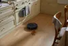 A black robot vacuum cleaner is cleaning up a spill on a wooden floor in a kitchen. Produktbild-Vorschau