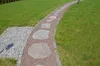 A winding garden path made of large stepping stones and red gravel, bordered by a cobblestone path and lush green grass. Produktbild-Vorschau